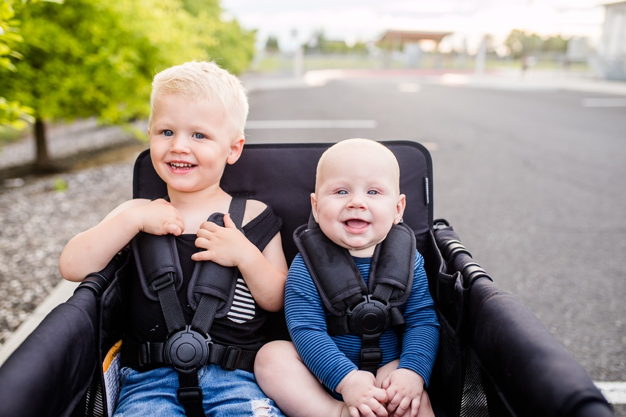 Two children smiling while buckled into 5-point harnesses within a W4 Luxe Pro stroller wagon.