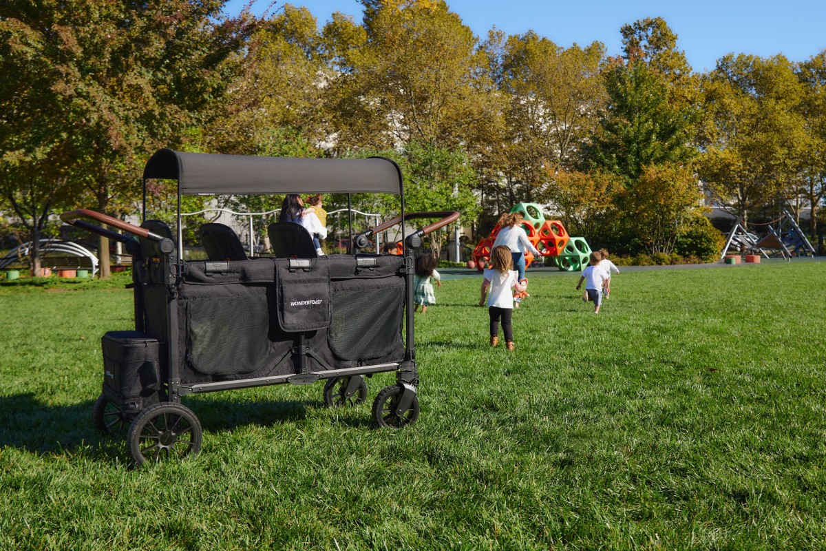 An empty W6 Luxe Pro stroller wagon at a park. Children can be seen running and playing in the grass.